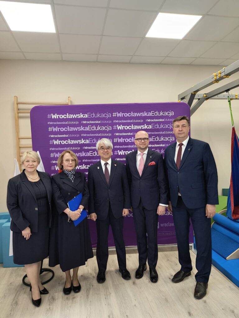 Grupa pięciu osób pozuje do zdjęcia na tle fioletowej ścianki z napisem "#WrocławskaEdukacja". W centrum stoi Jego Ekscelencja Akira Kono – Ambasador Japonii w Polsce, w towarzystwie przedstawicieli władz lokalnych i oświatowych. Uroczyste spotkanie odbywa się w placówce edukacyjnej, co podkreślają obecne elementy wyposażenia, takie jak drabinki i sprzęt terapeutyczny.